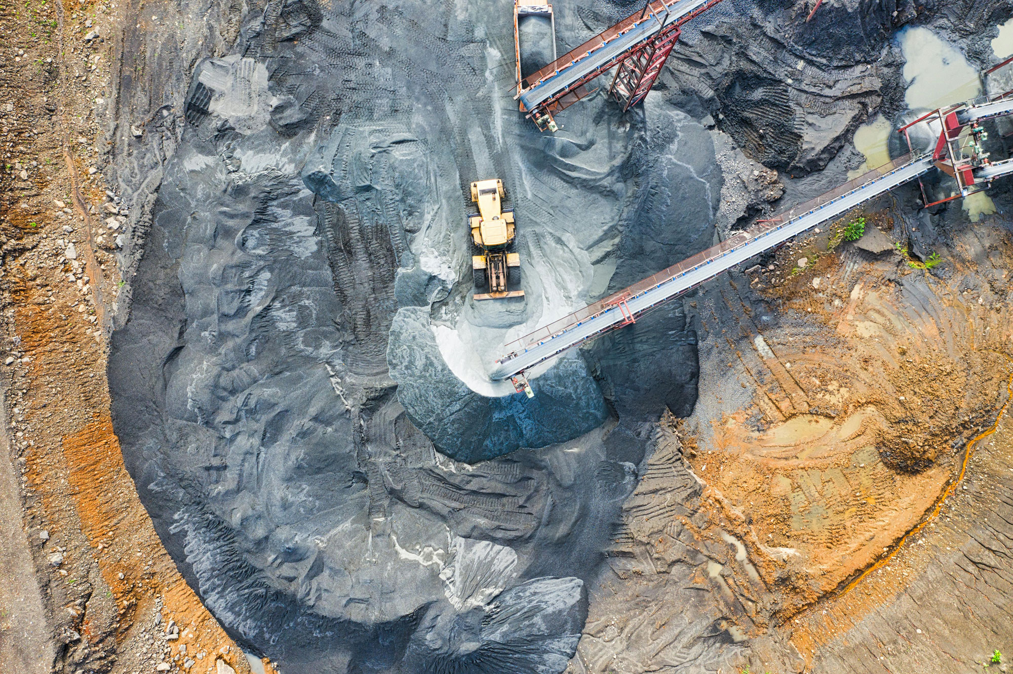Aerial view of a large mining operation with heavy machinery. A dump truck is being loaded with gray material from a conveyor belt. Surrounding landscape shows earthy tones with visible rock and soil layers.