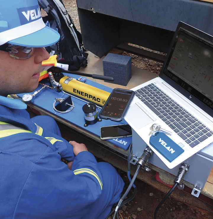 A person wearing safety gear and a helmet works with hydraulic equipment labeled "Enerpac" on a workspace bench. The person monitors data on a laptop, with a smartphone displaying a timer and various tools nearby. The name "Velan" is branded on their gear and tools.