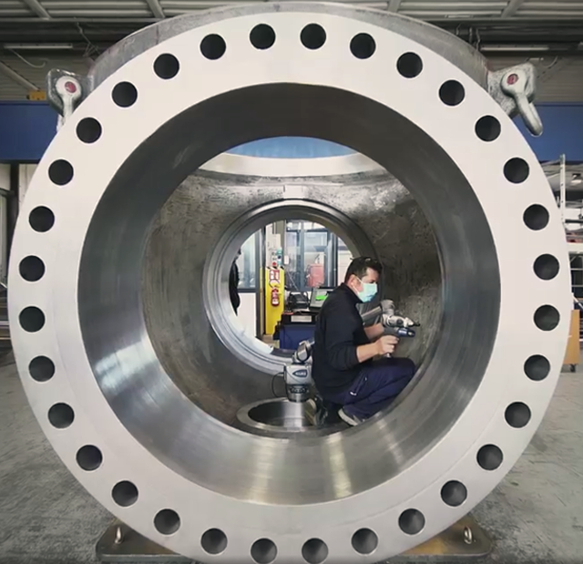 A worker in a protective mask is inside a large, cylindrical industrial pipe with multiple bolt holes around its rim. He is operating a handheld device for inspection or maintenance. The workshop setting includes machinery and industrial equipment in the background.